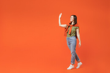 Full body side view young smiling happy woman 20s wearing khaki t-shirt tied sweater on shoulders walk go stroolling waving hand isolated on plain orange background studio. People lifestyle concept.
