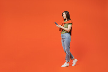 Fototapeta na wymiar Full body side view young smiling happy woman 20s wear khaki t-shirt tied sweater on shoulders hold in hand use mobile cell phone walk go isolated on plain orange background. People lifestyle concept.