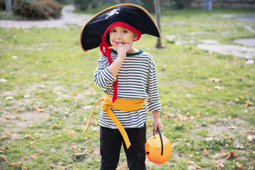 Treat or trick! Outdoor shot of little boy in pirate carnival outfit enjoying candy, eating lollipop standing in the park with pumpkin basket in his hand.