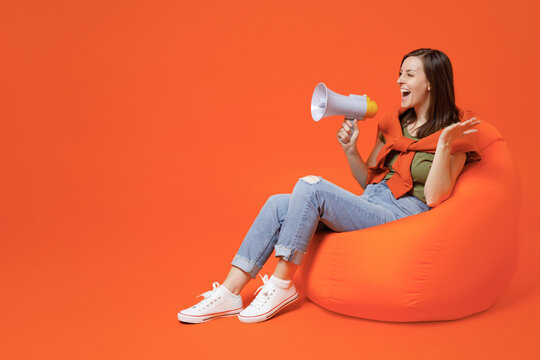 Full body young expressive happy woman 20s wear khaki t-shirt sweater sit in bag chair hold scream in megaphone announces discounts sale Hurry up spread hands arms isolated on plain orange background.