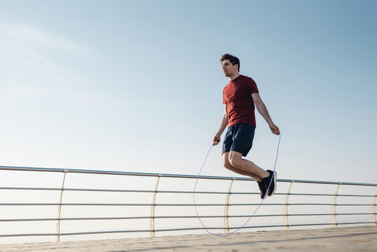 Full body young strong sporty toned fit sportsman man 20s in sports clothes jumping on skipping rope warm up training at sunrise sun dawn over sea beach outdoor on pier seaside in summer day morning