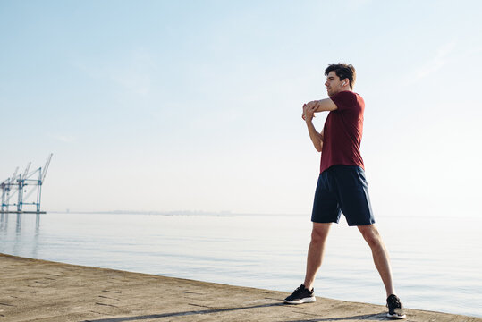Full body young strong sporty athletic toned fit sportsman man wear sports clothes do hand exercises warm up training at sunrise sun dawn over sea beach outdoor on pier seaside in summer day morning.
