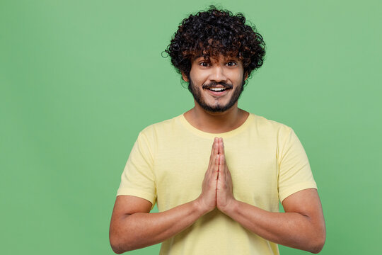 Young smiling happy Indian man 20s in basic yellow t-shirt hold hands folded in prayer gesture, begging about something isolated on plain pastel light green background studio People lifestyle concept.