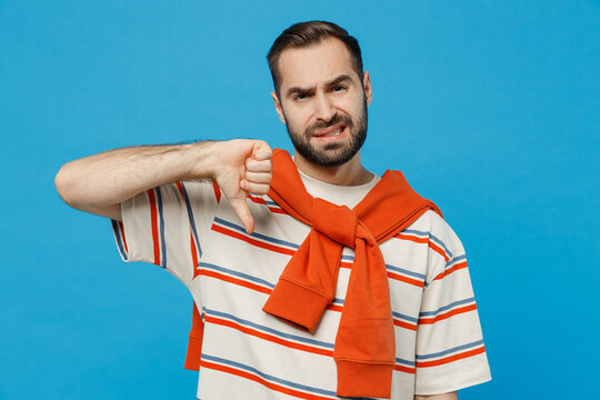 Young sad displeased dissatisfied caucasian man 20s in orange striped t-shirt look camera show thumb down dislike gesture isolated on plain blue background studio portrait. People lifestyle concept