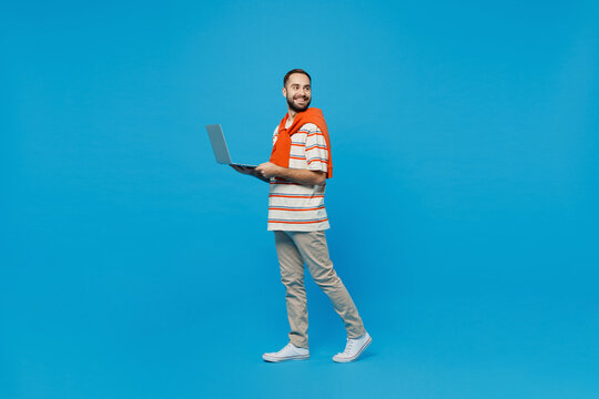 Full body young smiling fun happy man 20s in orange striped t-shirt looking camera hold use work on laptop pc computer look aside on workspace area isolated on plain blue background studio portrait