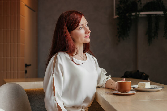 Smiling mid age woman with red hair hair sitting in cafe and drinking coffee looking away indoors