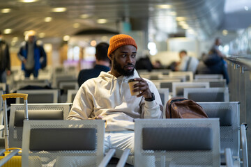 Young African American traveler man drinking coffee and eating sandwich while waiting for flying at airport terminal. Trendy black hipster guy satisfies hunger with a coffee and sandwich. 