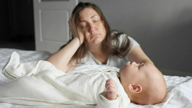 Tired mother sleeps sitting on floor leaning on bed with crying baby lying on bed white sheets. Newborn girl tries to draw attention of mother by crying
