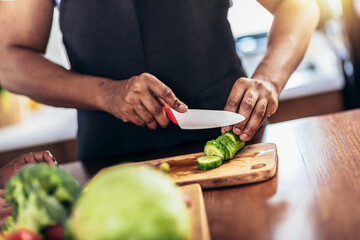 Close up shoot of black man holding sharp knife cutting cucumbers  on wooden board, making fresh...