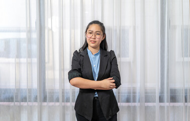 Little entrepreneur Asian eyeglasses woman with confident pose in black suit stands in front of window and curtain.