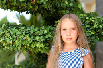portrait face of candid happy little kid girl of eight years old with long blond hair and green eyes on background of green plants during a summer vacation travel. gen z mental health concept