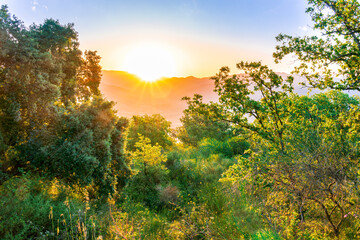 amazing highland landscape with scenic view from mountain with green branches and leaves on sides to a valley town with majectic mountains and scenic cloudy sunset on background