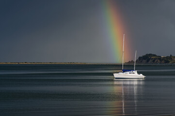 A rainbow appears to fall onto a yacht on a calm ocean