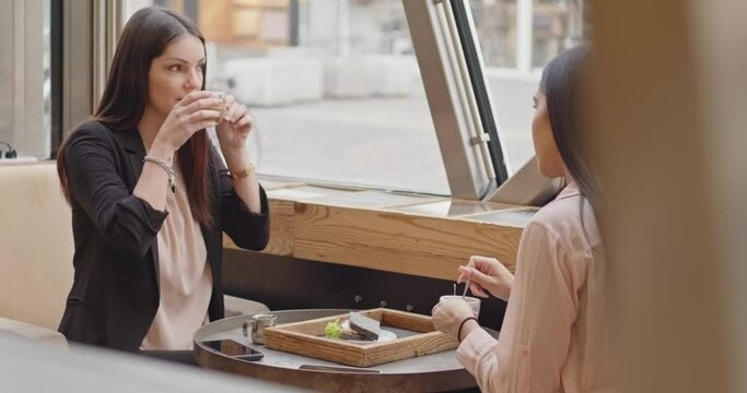 Back view of two adult business women sitting and talking in a bar. Having breakfast in a cafe, two multi-cultural friends are eating, talking, smiling and laughing