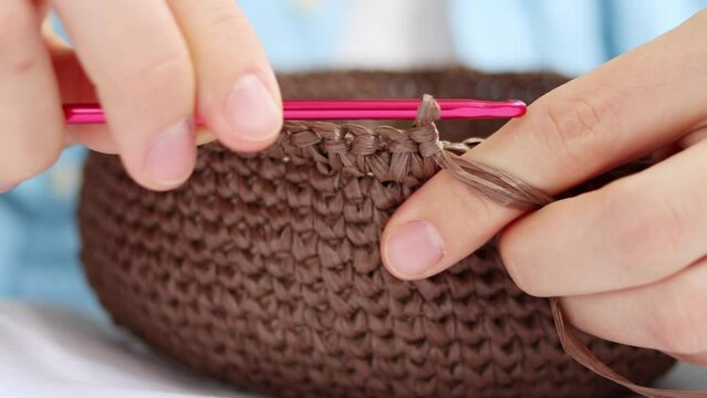 Front view of female hands knit crocheting a decorative brown basket using pink hook and raffia yarn close up. Creation of handmade accessories from natural materials, hobby. High quality 4k footage