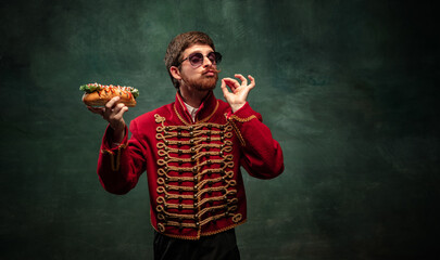 Young man in suit as royal hussar tasting big hot-dog isolated on dark green background. Retro style, comparison of eras concept.