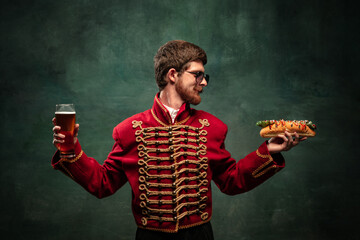 Young man in suit as royal hussar tasting big hot-dog isolated on dark green background. Retro style, comparison of eras concept.