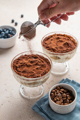 Traditional Italian dessert tiramisu in glass. Individual homemade layered cake in cup. Female hand sprinkles cocoa powder over tiramisu. Woman pours cocoa powder over cake. Selective focus.