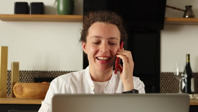 Beautiful young woman customer making mobile call confirming online website shopping order delivery concept. Talking on phone looking at laptop sitting in kitchen at home, wearing white clothes