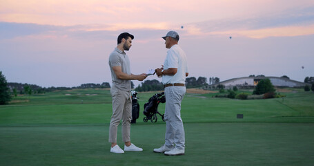 Two businessmen stand outside on golf course. Sportsmen sign paper on field.