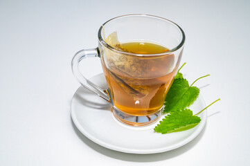 Nettle herbal tea in a transparent cup, with a sachet immersed in water, and a white saucer with nettle leaves next to it. Healing infusion of nettle. 
