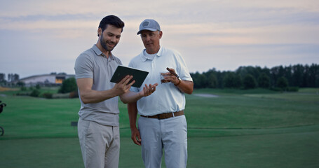 Two golfers search tablet on field landscape. Happy men chatting online outside.