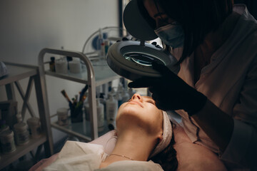 A beautician wearing medical gloves cleans the pores on the face using special equipment while...