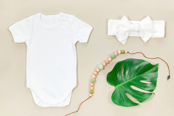 White bodysuit for baby mockup in eco style. Mockup of baby clothes on a beige background with teether beads, headband and a tropical leaf of monstera.