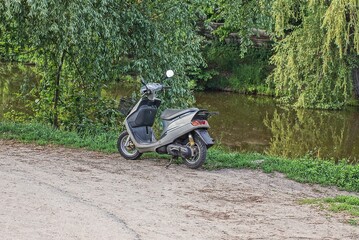one gray moped stands by the road in green grass on the shore of a lake with brown water