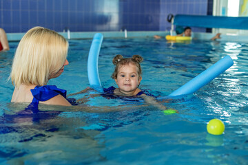 Mom with beautiful curly toddler girl play in swimming pool. Foam stick for swimming.