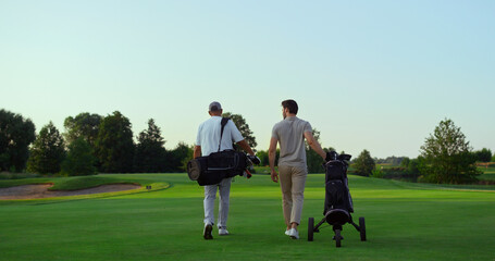 Rich golf players walking on field. Two active sportsmen carry equipment clubs.