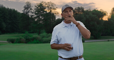 Wealthy elderly smoke cigar on golf field. Old guy posing looking camera outside