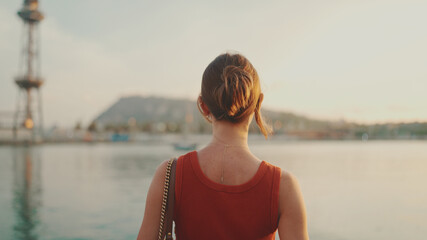 Girl stands on the embankment and looks at the bay. Young woman looks at the sea in the morning time. Back view