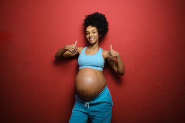 Portrait of fit sucessful black pregnant woman on fitness sportswear doing thumbs up gesture against red background. Pregnancy healthy lifestyle and success concept.