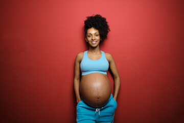 Portrait of healthy relaxed black pregnant woman on fitness sportswear against red background.