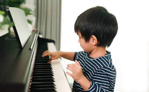 Close up of asian toddler boy is practice playing piano at home and smiling with happiness moment, concept of learning, art, steam, musical, mental health, homeschool, skill, ability concept.