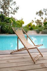 Empty bamboo chair next to swimming pool in private residential area. Wooden floor and green area in the background.
