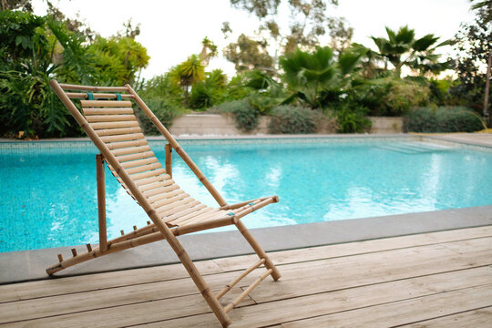 Empty bamboo chair next to swimming pool in private residential area. Wooden floor and green area in the background.
