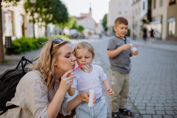 Young mother playing with her kids, blowing bubbles in city street in summer.