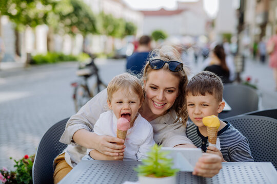 Young mother taking selfie with her kids eating ice-cream in cafe outdoors in street in summer.