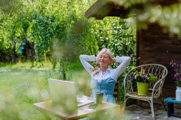 Senior woman with laptop and coffee sitting on terrace in summer, resting.