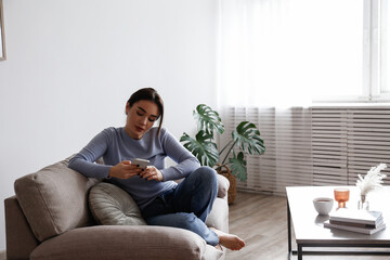 Fototapeta premium Portrait of young brunette woman with concentrated facial expression sitting on the couch typing on the phone. Joyful female having a video call. Background, copy space, close up.