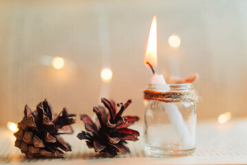 burning candle in a glass jar, cones on the background of an old open book. Festive bokeh lights in...
