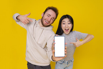 Caucasian guy and asian girl show thumbs up excited holding smartphone with blank white screen, mobile app advertisement isolated on yellow background. Product placement. Great offer