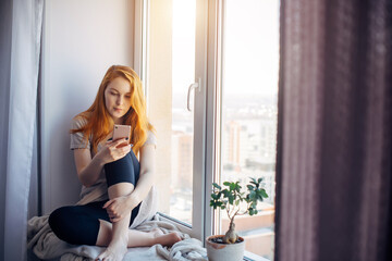 Obraz na płótnie Canvas Beautiful red-haired woman sitting on the windowsill in a cozy city apartment. Young girl spends her leisure time using smartphone.