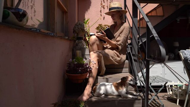 writting notes sitting on wooden stairs in contryside style yard with small dog. Relaxed summer day mood. Woman in linen natural dress and straw dress. Small dog lies at the feet. Video footage