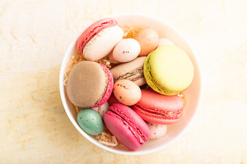 Different colors macaroons and chocolate eggs in ceramic bowl on beige concrete background. top view, close up.