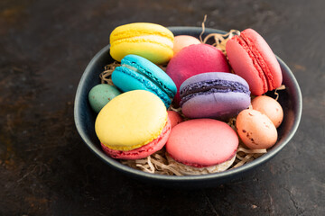 Different colors macaroons and chocolate eggs in ceramic bowl on black concrete background. side view, close up.