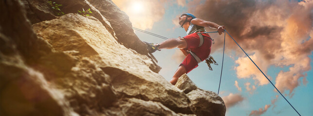 Fototapeta Muscular climber man in protective helmet abseiling from cliff rock wall using rope Belay device and climbing harness on evening sunset sky background. Active extreme sports time spending concept. obraz