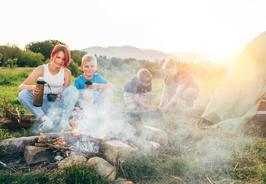 Group of smiling kids near a smoky campfire drinking tea from a thermos, two brothers set up the green tent. Happy family outdoor picnic camping activities concept